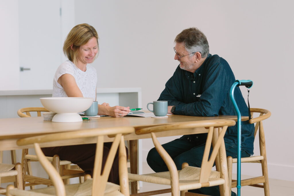 An older man with a walking stick and a therapist sitting at a dining table at home, going through his notes together